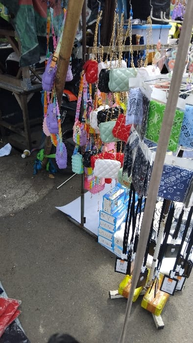 The image shows a street vendor's stall with various colorful purses and small bags hanging on a metal rack. The purses are made of different materials, including leather and fabric, and come in various shapes and sizes. They are attached to chains and hung from a metal rack, with multiple price tags hanging behind them. The image captures a view from the side, looking down at the items. It is a street stall in Kecamatan Margahayu, Indonesia (latitude: -6.963, longitude: 107.575).  The stall is set up on a concrete street, with some debris in the background.