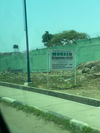 This image captures a slightly tilted view of a sign for "Mubeen International College" in Bauchi, Nigeria. The sign is white with green text and Arabic lettering at the top. Below the college name, it lists the school's motto as "Knowledge is Light" and provides details about "Nursery, Primary and Islamic School," "Adult Arabic and Islamic Studies," and "House Wife Juta Aure Maiic and Islamic Studies." An arrow points to the right, indicating a direction.
The sign is positioned alongside a green, textured wall, possibly a building or boundary, with barbed wire running along its top. To the left of the sign, a large tree with dense foliage is visible, its branches reaching towards the sky. Behind the wall, a rocky, overgrown area suggests a natural or undeveloped landscape.
In the background, a metal structure with water tanks is visible, possibly a water tower or part of a larger facility. The sky is a clear, pale blue, indicating daytime. The overall scene appears to be outdoors, by a road or pathway, with the green wall and surrounding vegetation suggesting a somewhat rural or semi-urban setting within Bauchi. The image is taken from a moving vehicle, as evidenced by the blurred foreground and the reflection on what appears to be a car window.