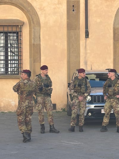 The image, set in Siena, Italy, depicts a daytime scene featuring four soldiers in camouflage uniforms standing outdoors on a paved surface. They are positioned in front of a light-colored historical building and a dark vehicle.
The four male soldiers are uniformly dressed in military camouflage fatigues, tactical vests with various pouches, and distinct dark red berets. They also wear dark combat boots. Their stances suggest they are on duty, possibly guarding the location or on patrol, with neutral, vigilant expressions, looking straight ahead or slightly upwards.
Behind the soldiers, a dark-colored SUV or jeep is partially visible. The front of the vehicle shows part of its grille and headlights. A portion of its license plate is visible, displaying the characters "...EJ 88".
The background consists of an old, light-tan stucco building with prominent architectural features. These include a large stone archway on the left side of the frame, decorative stone trim along the building's facade, and a dark barred window on the lower left. A dark pipe or drainpipe is also visible against the wall. The ground beneath the soldiers and the vehicle is a grey paved surface, possibly cobblestone or asphalt.
The lighting is bright and even, indicating a clear day, likely late morning or early afternoon. The overall atmosphere conveys a sense of security and military presence within a historical urban setting.