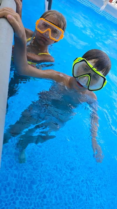 Two children, one wearing orange goggles and the other wearing yellow and black goggles, are in a bright blue swimming pool. The child in orange goggles is in the foreground, looking towards the camera with a neutral expression, and is holding onto the side of the pool. The child in yellow and black goggles is in the background, facing away from the camera, and appears to be moving through the water. The water is clear and the bottom of the pool is visible, showing a patterned texture. The lighting suggests it is daytime, likely outdoors.