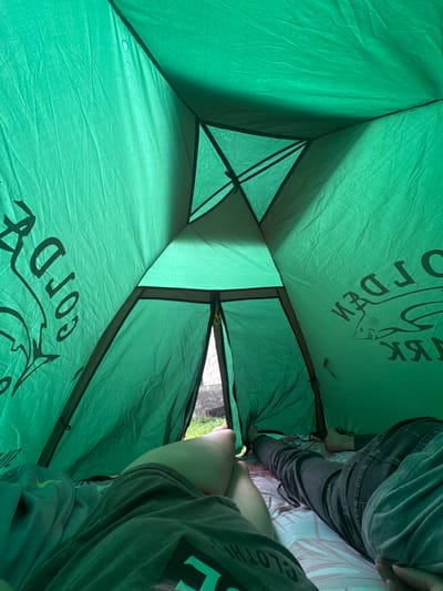 The image shows the interior of a green tent. Two people are lying down inside the tent, on sleeping bags or mats. Their legs are visible. One person's legs are closer to the camera and are covered by a green t-shirt. The other person's legs are further away and are covered by dark pants. The tent is made of waterproof material and has condensation droplets on the inside. There is text and a shark graphic printed on the tent fabric, appearing to be a brand name. The entrance of the tent is partially open, revealing some greenery and a building outside. The location is stated to be in the city of Узюково, Russia.