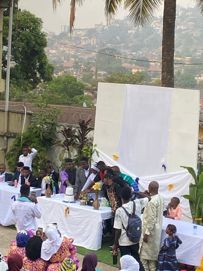 An outdoor social gathering is depicted on an elevated terrace or balcony, overlooking a sprawling, densely populated hilly cityscape, characteristic of Freetown, Sierra Leone. The scene is under soft, diffused lighting, suggesting late afternoon or an overcast day.

Numerous individuals, mostly adults with a few children, are present, seated and standing around white-covered rectangular tables. The attire is diverse, including traditional African garments such as men's long kaftans (some in light tones, others in olive green) and women's vibrant patterned dresses, mixed with more casual clothing. Many men are wearing white prayer caps or kufis.

The tables are draped with white tablecloths and hold bottled water, plastic cups, and decorative elements. A small white two-tier item with blue accents, possibly a cake or centerpiece, is visible, along with arrangements of yellow artificial flowers. White fabric panels serve as a backdrop or overhead covering, decorated with small yellow and blue ribbons or details. Lush tropical vegetation, including a large palm tree trunk and fronds on the left, and banana leaves behind one table, contributes to the outdoor setting.

Attendees are engaged in various activities, including socializing and interacting with items on the tables; some individuals are looking at their mobile phones. The overall atmosphere appears relaxed and communal, indicative of a special occasion or social event. The background reveals numerous buildings scattered across the hazy hillsides under a pale sky. No legible text is visible on any objects or clothing.