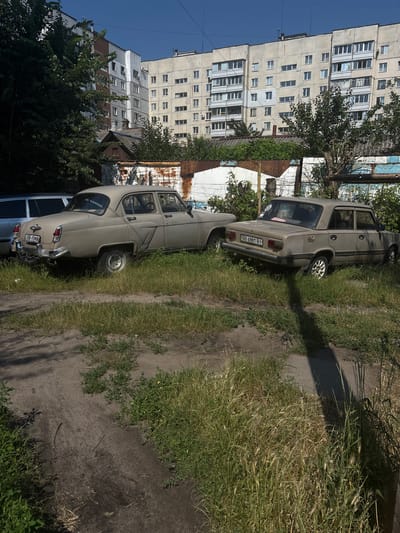 The image depicts a street scene in Cherkasy, Ukraine, under a clear sky on a sunny day. The perspective is rotated 90 degrees counterclockwise. In the foreground, two vintage cars are parked along a dirt and grass path. The closest car is a light beige sedan with chrome accents and a visible license plate reading "08 6984 BI". Behind it, another car, also a light beige sedan, is partially visible. Overgrown grass and weeds line the path, with shadows cast by unseen objects suggesting mid-morning or afternoon sun.
In the background, a tall, grey, multi-story apartment building dominates the left side of the frame. Its facade is characterized by numerous windows and balconies. Adjacent to it, another, slightly shorter apartment building with a mix of grey and brick-colored sections is visible. Trees and foliage are interspersed between the buildings and the parked cars, adding a touch of green to the urban environment. A weathered, corrugated metal fence or structure is also present, contributing to the scene's slightly dilapidated but lived-in feel. The overall impression is of a typical residential area in an Eastern European city.