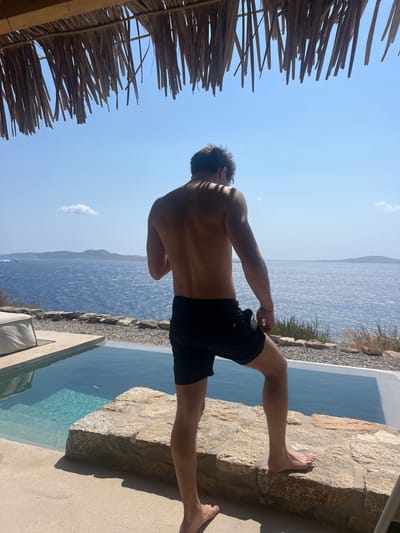 An outdoor scene in Agios Ioannis Diakoftis, Greece, depicts a single male subject positioned with his back to the viewer, facing a large body of water. The subject, shirtless and wearing black swim shorts, stands barefoot with his left foot elevated on a natural stone retaining wall that runs across the mid-ground. Shadows from an overhead thatched or straw roof structure fall horizontally across his upper back and head.
To the subject's left and in the foreground, an infinity-edge swimming pool with light blue water is visible, bordered by a concrete patio. A light-colored outdoor lounge cushion is partially visible on the far left. Beyond the stone wall, the environment transitions to sparse, dry Mediterranean vegetation. The expansive deep blue Aegean Sea dominates the background, exhibiting ripples on its surface. Distant landmasses, characteristic of Cycladic islands, are discernible on the horizon. The sky above is clear and bright blue, with one small white cloud visible in the upper left quadrant. The scene is illuminated by direct, bright sunlight.