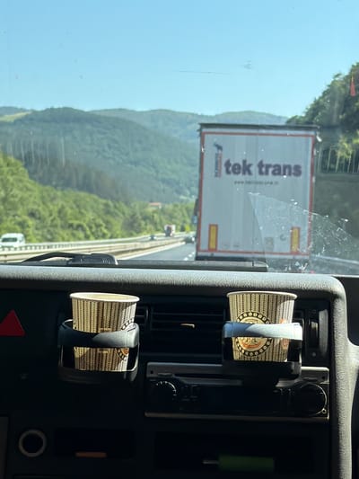 An interior view from a vehicle shows a dashboard with two paper coffee cups in cup holders. The cups have a repeating pattern of arrows and coffee beans. Beyond the dashboard, a highway is visible with traffic, including a white van and a large white truck with "tek trans" written on it. The truck has red and yellow accents. Mountains covered in green vegetation are in the background under a clear blue sky. The scene is likely on a road near Churek, Bulgaria.