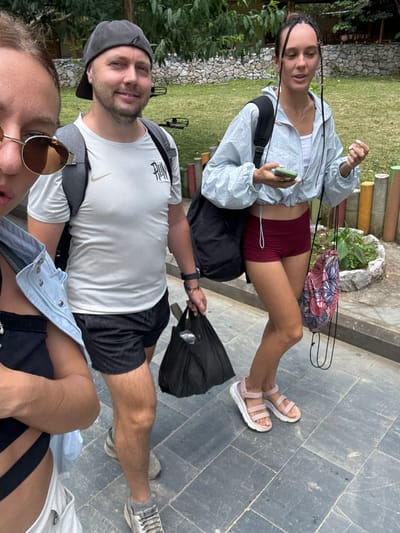 Three people stand on a paved surface, likely in Mèo Vạc Commune, Vietnam. The woman in the foreground wears sunglasses and a blue denim shirt over a black top. A man next to her is dressed in a white Nike T-shirt, black shorts, a gray cap, and sneakers, holding a black plastic bag. A second woman, standing farther back, is wearing a light blue jacket, red shorts, and pink sandals, holding a phone. Backpacks are visible on the backs of both women and the man. The background includes grass and a low wooden fence.