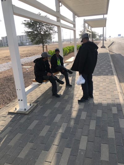 Three adult males are observed under a white metal shelter with a corrugated roof on a paved walkway in Hanak, Saudi Arabia, during daytime. Two males are seated on a light-colored bench within the shelter, facing each other. Both wear dark jackets, dark trousers, dark shoes, and grey baseball caps; the male on the right also wears a yellow-green high-visibility garment under his jacket and is smiling. A third male stands with his back partially to the viewer, facing the seated individuals, similarly dressed, and holds a white plastic bag in his left hand. The paved walkway consists of rectangular grey and darker grey interlocking pavers, bordered by a gravel area to the left and an asphalt road with a curb to the right. Long shadows are cast across the pavement from the shelter and individuals. In the background, sparse trees and low green bushes are visible on arid ground, with large, light-colored industrial-style buildings under a hazy sky in the distance.