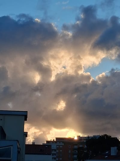 The image shows a section of the sky filled with dark, billowy clouds with light shining through gaps in the clouds. Buildings are visible on the right side of the image, one with a dark facade. The image may be taken from Figueira da Foz, Portugal, but it is hard to determine. There are no people, subjects or actions in the image.