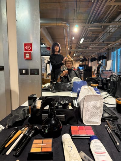 The image shows a table with various makeup products and tools, including brushes, palettes of blush, eye shadow and lipstick, cream tubes, and cosmetic bags. The table is covered in black fabric. In the background, a mirror reflects the interior of a building, including industrial-style ceilings with pipes and metal beams, as well as people including two women. The photo appears to be taken in a backstage or preparation area, likely in a studio or production space. A fire alarm system, including an alarm with a starburst-type image and an outlet, can be seen on the wall in the mirror's reflection. There is no information in the photo that specifies the city or state. The location is Preara, Italy is not stated in the picture.