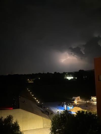 This is a nighttime photograph taken from an elevated perspective, likely a balcony or window, overlooking a landscape that includes urban elements and natural features. The sky is dark and cloudy, illuminated by a prominent lightning strike that forks across the upper portion of the image. Below the stormy sky, a dense line of trees stretches horizontally across the middle ground, obscuring much of the terrain behind it.

In the foreground, to the right, a portion of an orange building with a window is visible. To the left, a large, pale, sloped structure dominates the lower part of the frame. This structure appears to be man-made and illuminated by lights, possibly a retaining wall or part of a building. A red light is visible on the far left of this structure.

In the middle ground, beyond the trees, a few scattered lights indicate buildings or infrastructure. Closer to the foreground, and nestled within the landscape, there are several temporary structures, including illuminated tents and food trucks. One food truck, white with a yellow illuminated sign, is visible. People are present in this area, appearing to be gathered around the tents and food vendors. A series of small lights are arranged in a line along the edge of the sloped structure in the foreground.

The overall scene depicts a stormy night with a dramatic lightning display over a partially visible urban or suburban environment, possibly a park or outdoor event space, given the presence of food trucks and gathering people. The location is identified as Glattbrugg, Switzerland.