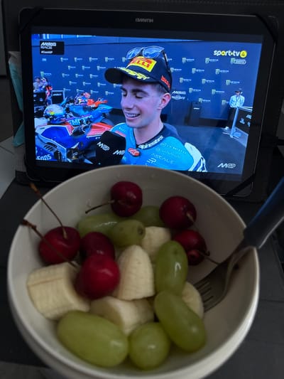 In the city of Almada, Portugal, a tablet displays a MotoGP race broadcast, featuring a young male rider in a helmet and racing suit smiling. To the left of the rider on the screen is a "Moto2 RACE #CzechGP" logo, and to the right is a "sport.tv4" logo. In the foreground, a white bowl contains a fruit salad with sliced bananas, green grapes, and cherries. A fork rests on the edge of the bowl, with its tines partially submerged in the fruit. The tablet is set against a dark surface, possibly a table or stand.