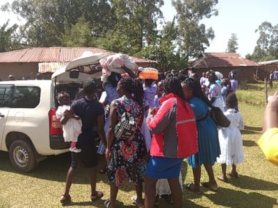 A gathering of people surround a white van with its trunk open, as items are being unloaded. One person holds a young child. The group appears to be composed of women, many wearing white dresses, suggesting a religious or special event. In the background are buildings with corrugated metal roofs, situated among green trees and grass, potentially indicating a rural or outdoor setting.