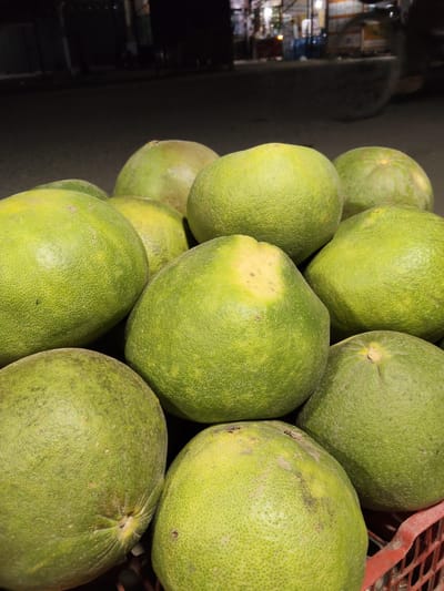 A close-up, low-angle shot captures a pile of green pomelos, or "jeruk bali," arranged in a red plastic crate. The fruits, with their textured rinds, vary slightly in shade from bright green to yellowish-green, with some showing subtle brown blemishes or lighter patches. The background is dark and out of focus, hinting at an outdoor setting, possibly a market at night, with blurred lights suggesting street lamps or shop displays. The lighting is dim, highlighting the round forms of the pomelos and creating a slightly moody atmosphere. The arrangement is natural, with the fruits nestled together, showcasing their abundance. There are no people visible, and the focus is solely on the produce. The overall impression is of fresh fruit, likely ready for sale or consumption, presented in a simple, unadorned manner. The setting, given the context of Peurupok, Indonesia, suggests a local market scene. The time of day appears to be evening or night due to the low light conditions and the illuminated background elements.