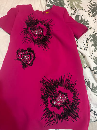 An overhead shot depicts a fuchsia-colored, short-sleeved dress laid flat on a patterned surface in the city of Vladaya, Bulgaria. The dress features a simple scoop neckline and is made of a smooth, structured fabric. Three distinct, abstract designs are visible on the front of the garment. Each design is rendered in black lines radiating outwards, resembling a spiky floral or starburst pattern. The center of each design is adorned with a cluster of small, clear, faceted rhinestones and tiny silver beads. One large design is centered on the upper chest, a smaller design is positioned below and to its left, and another large design is situated towards the lower right portion of the dress. The dress is laid upon a textile background featuring an organic pattern of large olive green leaf-like shapes and scattered black polka dots on a white or cream base, consistent with a duvet cover or sheet. A plain white textile surface is visible along the left edge of the frame.