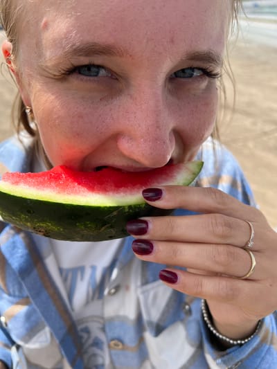 In this close-up, vertically oriented image, a person is shown eating a slice of watermelon. The individual's face is partially visible from a low angle, with their eyes looking upwards and to the left. They have fair skin with freckles and blue eyes. Their mouth is open, taking a bite of the watermelon, which has a bright red interior and a green rind.
The person is wearing a blue and white plaid shirt. Their right hand is visible, holding the watermelon slice. The hand has dark red nail polish, and there are two rings on the ring finger, one appearing to be a simple band and the other a thin band with a small gem.
The background is blurred, but appears to be an outdoor setting, possibly a beach or picnic area, with light-colored sand or fabric. The overall lighting suggests it is daytime, likely sunny, given the brightness. The scene evokes a sense of casual enjoyment and refreshment on a warm day. No text is visible in the image. The location is identified as Alexandrovka, Russia.