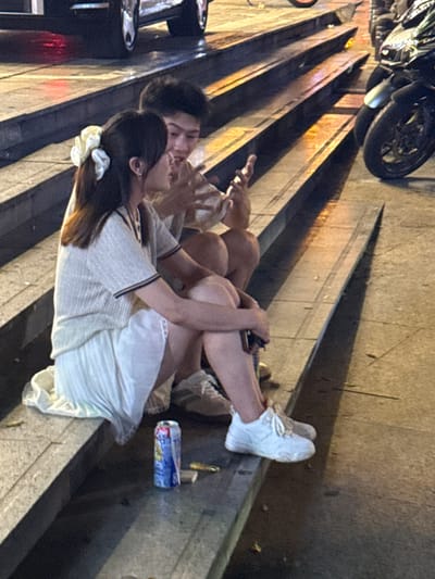 A young Asian couple sits on concrete steps in what appears to be a pedestrian area, possibly a park or plaza, at night in Longta, China. The woman, on the left, has dark hair adorned with a white scrunchie and wears a light-colored, short-sleeved ribbed sweater and a white pleated skirt. The man, on the right, has dark hair and wears a light-colored shirt and shorts. They are seated close together, and the woman appears to be looking at the man, who is gesturing with his hand. A blue and red canned beverage and a golden object are on the step in front of the woman. In the background, a motorcycle is parked, and a car is visible further back, suggesting an urban setting. The lighting suggests artificial streetlights or ambient light from nearby establishments.