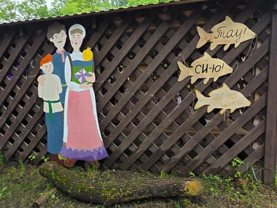 This media file shows an outdoor scene with a wooden fence adorned with painted figures and wooden fish shapes. On the left, there are cutout figures of a family: a father with a mustache and blue shirt, a mother in traditional dress holding a baby, and a young boy with red hair. They are painted in a folk art style.
Attached to the fence are three wooden fish cutouts, each with handwritten text in Russian. The top fish says "спасибо! Грay! спасибо!". The middle fish reads "ЕШЬ-ПЕЙ! СИ-Ю! ЕШЬ - ПЕЙ!". The bottom fish has the text "точ-точ сиськы!". The fence itself is made of dark brown wooden slats arranged in a diagonal lattice pattern. Behind the fence, some structure is visible, possibly a gazebo or shed, with patterned fabric hanging inside.
The scene is set outdoors, with green grass and weeds growing at the base of the fence, and a fallen moss-covered log in the foreground. Trees with green foliage are visible above the fence, suggesting it is daytime and likely spring or summer. The sky is overcast, indicating potentially cloudy weather. The overall impression is rustic and whimsical, with the folk art and handwritten signs adding a touch of local charm. The location context is identified as "Старые Быги, Russia".