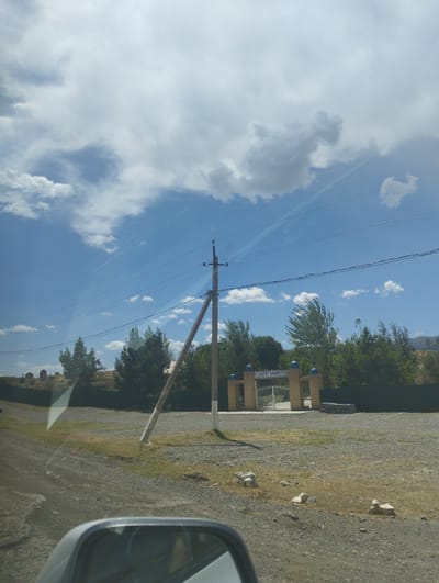 The image shows an outdoor scene with a clear sky and some clouds. In the foreground, the edge of a car's side mirror is visible. A dirt road leads towards a gateway with an inscription that appears to read "ILONCHI QABRISTONI". The gateway is constructed of brick and has blue domed finials. Beyond the gateway, there are trees and a dark fence. To the left of the gateway, there are gravestones on a hillside. A wooden utility pole with power lines is also visible in the scene. The environment appears to be rural, possibly in the city of Asmansay, Uzbekistan.