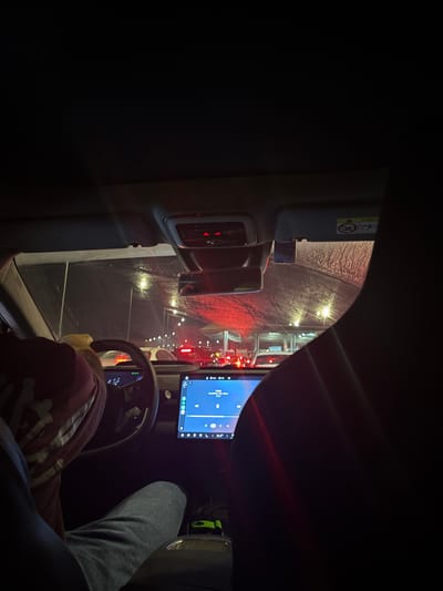 The image shows the interior of a car at night, appearing to be in traffic. The perspective is from the passenger seat, looking forward. The windshield is covered in what appears to be condensation or frost, with some streetlights and red taillights visible through it, suggesting an urban environment. A person's left arm and shoulder are visible in the foreground, wearing a red sweatshirt with white graphics. The steering wheel and dashboard are also visible. The car's central touchscreen displays a media interface with song information and playback controls. Red traffic lights are prominent in the distance, illuminating the scene. Based on the provided crops and context, the location is identified as Modi'in Illit, Palestinian Territories.