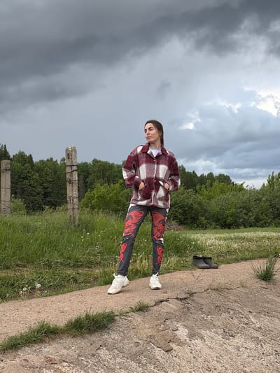 A young woman stands outdoors with a grey, overcast sky above. She wears a maroon and white plaid jacket over a white shirt, black leggings with a red design, and white sneakers. The environment includes trees and grass with dandelions. A cracked concrete edge is visible, bordering the grass. Two black shoes are discarded further along the concrete edge. In the distance are the remnants of broken concrete structures entwined with barbed wire. The location is Nizhnii Vishur, Russia.