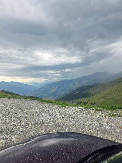 An overcast sky looms over a mountainous landscape. In the foreground, the hood of a dark-colored vehicle is visible, speckled with dust. Beyond the vehicle, a gravel road curves into the distance, bordered by green grass and wildflowers. The road leads to a panoramic view of a valley with a town nestled within, surrounded by rolling hills and majestic mountains that recede into the hazy distance. The scene appears to be a scenic overlook in the vicinity of Séez, France. There are no people visible in the image.