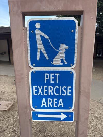 A close-up, eye-level shot shows a brown metal post holding three blue and white signs. The top sign depicts a white silhouette of a person walking a dog on a leash, with a white border and reflective pattern. Below it, a larger sign reads "PET EXERCISE AREA" in white, reflective capital letters on a blue background, also with a white border and reflective pattern. The bottom sign is a smaller blue rectangle with a white arrow pointing to the right, bordered by white and a reflective pattern. The signs are secured to the post with visible rivets. The background is out of focus but appears to be a sandy or gravelly outdoor area with some sparse vegetation and a building in the distance. The scene is outdoors, likely in a public park or recreational area.