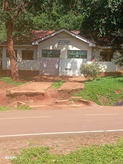 The image captures a public washroom facility situated amidst lush green foliage and trees, likely in Nkomo ward, Kenya, during the daytime. The single-story building features light-colored walls and a reddish-brown tiled roof. Prominently displayed on the front gable is a sign that reads "WASH ROOMS." To the left, above an entrance, "LADIES" is visible, while to the right, another sign indicates "GENTS." A person wearing a red top is partially visible standing inside the "GENTS" section.
The building has multiple rectangular windows with green frames. A section of the wall between the main entrance and the "LADIES" side is covered in light-colored tiles. A dirt path, appearing somewhat eroded and tiered, leads up to the building from a paved road in the foreground. The road has a single white dividing line and appears to be made of reddish-brown asphalt. Beyond the path, the ground is covered with sparse green vegetation. The background is dominated by dense, dark green trees and foliage, suggesting a natural or semi-forested environment. The lighting suggests a bright, clear day. A "REDMI 13c" watermark is visible in the bottom left corner, indicating the device used to capture the photo.