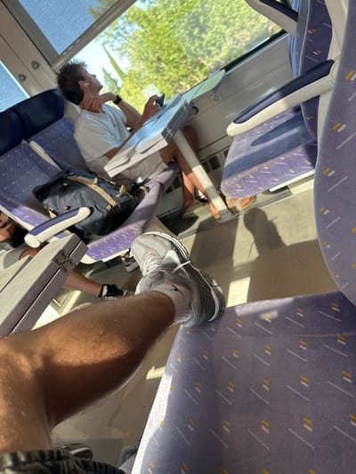 The image depicts the interior of a train carriage, likely in or near Salses-le-Château, France, given the sunny, tree-lined scenery visible through the window. In the foreground, a person's legs are visible, with one leg extended forward wearing a grey athletic shoe and white socks. The seat in the foreground is upholstered in a purple fabric with a geometric pattern of white and yellow lines. Adjacent to this seat, another seat is occupied by a person wearing a white t-shirt and grey shorts, looking out the window. This person is holding a mobile phone in their right hand and has wireless earbuds in their ears. A navy blue backpack rests on the seat next to them. A fold-out tray table is positioned in front of the person in the white t-shirt, with the mobile phone on it. Sunlight streams into the carriage, casting shadows on the floor and seats. The overall environment suggests a journey in progress, with passengers engaged in typical travel activities.