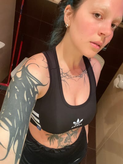 A fair-skinned person with light eyebrows and dyed blue hair is posing for a selfie in what appears to be a bathroom. The person is wearing a black Adidas sports bra and black shorts. The person has multiple tattoos on their chest, stomach, and arms. The bathroom features dark brown tiled floors, light brown walls, a toilet, and a toilet paper dispenser. The location is not discernible as being Villaviciosa, Spain, based on visual information.
