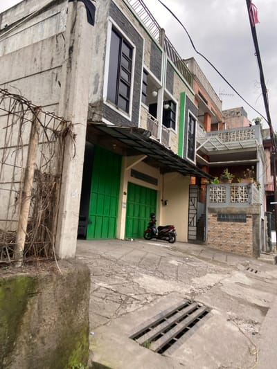 The image shows a narrow street in Margaasih, Indonesia (latitude: -6.961, longitude: 107.546), lined with two-story concrete buildings. The street slopes downhill, with a drainage grate visible in the foreground. A motorcycle is parked in front of a green garage door, and a tall concrete wall with a metal fence is visible on the left side of the image. An overhead power line runs along the buildings. There is no one visible in the image.