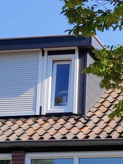 The image shows the upper facade of a modern house, likely in the city of Breda, Netherlands. The roof is covered with terracotta tiles arranged in a typical overlapping pattern. Above the roofline, there is a dark-colored eave and a section of white siding. A tall, narrow window with a white frame is visible, reflecting a blue sky and some green foliage. To the left of this window, a roller shutter is partially extended. A branch with green leaves from a tree extends into the upper right corner of the frame. The overall scene suggests a residential building on a clear day.