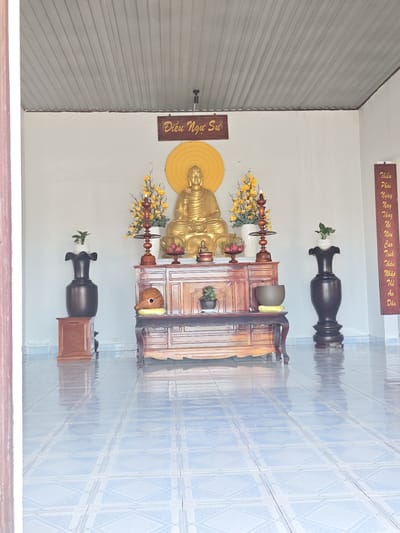 The image depicts an altar with a golden Buddha statue at its center, seated on a wooden cabinet. The altar is adorned with yellow floral arrangements and other decorative items. To the left of the Buddha statue, a tall, dark vase with a small plant sits on a wooden stand. To the right, another similar vase is present. A vertical inscription with Vietnamese text is visible on the far right wall. The floor is tiled with a blue and white pattern, and the ceiling appears to be made of corrugated metal. The scene is set indoors, likely within a temple or shrine, in the city of Phan Rang – Tháp Chàm, Vietnam. There are no people visible in the image.