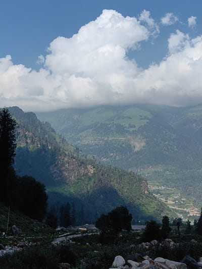 The image captures a panoramic view of a mountainous landscape near Palchan, India, under a partly cloudy sky. In the foreground, rough terrain with large rocks and dense vegetation dominates, with a dark silhouette of trees on the left side of the frame. A winding road or path is visible in the mid-ground, with a few scattered trees and rocky outcrops.
Further into the scene, steep, verdant mountains rise, covered in lush green trees and sparse rocky patches. Shadows cast by the clouds highlight the contours of the slopes. In the distance, the mountains recede, with layers of green hills and valleys. A small settlement or village with buildings is visible nestled on the slopes of the far mountains. A faint waterfall can be seen cascading down one of the distant peaks. The sky above is a vibrant blue, interspersed with large, fluffy white clouds. The overall impression is one of a serene and natural mountainous environment.