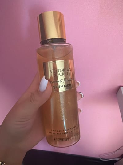 A hand with white nail polish holds a Victoria's Secret Coconut Passion Shimmer fragrance mist bottle against a pink background. The bottle is transparent, revealing a golden liquid with glitter. The label on the bottle reads "VICTORIA'S SECRET Coconut Passion SHIMMER" and the volume is indicated as "250 mL / 8.4 fl oz". The background appears to be a textured pink surface, possibly a wall or fabric. There are no identifiable landmarks or elements that suggest a specific location in Novi Iskar, Bulgaria. The lighting suggests an indoor setting.