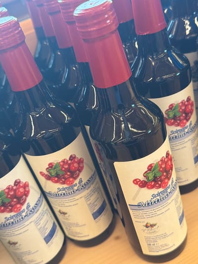 The image shows a close-up, angled view of several bottles of "Sciroppo di Mirtillo Rosso" (Red Blueberry Syrup) lined up on a wooden surface, likely a shelf. The bottles have dark red caps and clear glass, revealing the dark red liquid inside. Each bottle features a white label with an illustration of red berries and green leaves, along with text in Italian. The text on the labels includes "Sciroppo di Mirtillo Rosso," "INGREDIENTI: Zucchero, sciroppo di glucosio-destrosio, succo di mirtillo rosso, da succo concentrato 33% DA DILUIRE 1:7," and "Produttto e Confezionato da Azienda Agricola Dolomites Albert, Via Laretta N°14 - 33020 SAURIS (UD)." The location "Sauris (UD)" indicates that the product is from Sauris, Italy. The overall lighting is bright, with some glare on the bottles.