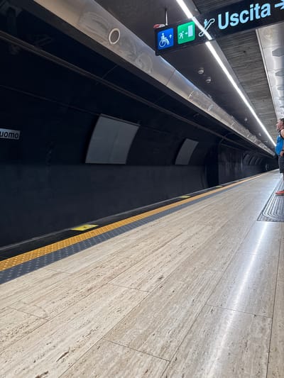 The image depicts a subway station platform in Naples, Italy. A sign overhead reads "Uscita," meaning "Exit" in Italian, with directional arrows and symbols indicating accessibility and stairs. The platform is made of light-colored tiled flooring, with a yellow and grey textured tactile paving strip along the edge. On the left side of the image, a dark wall features the word "Uomo" in white lettering, likely indicating a men's restroom or changing facility. The station appears to be modern and well-lit, with recessed lighting along the ceiling. A person, partially visible on the right, is standing on the platform, looking towards the tracks. The overall scene suggests a typical, functional public transportation environment.
