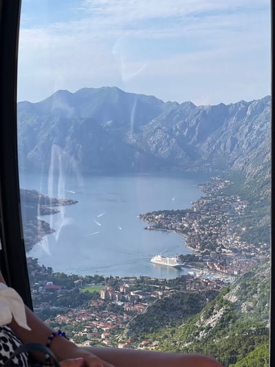 The image presents a high-angle view, likely from a cable car or elevated vantage point, overlooking a expansive bay and a densely settled coastal area identified as Mirac, Montenegro. In the immediate foreground, the dark frame of a window or cabin is visible on the left and right, with reflections on the glass. A person's right arm and shoulder are partially visible in the lower left, adorned with a blue beaded bracelet and a black-and-white patterned garment.

The central focus of the scene is a large, calm body of water, a bay or fjord, surrounded by steep, rugged mountains. Several small boats and speedboats are visible on the water, leaving white wakes. A prominent feature in the bay is a large, white multi-deck cruise ship, anchored or docked near the city's waterfront.

The city, referred to as Mirac, stretches along the coastline and extends up the lower slopes of the surrounding mountains. It consists of numerous buildings, predominantly with red-tiled roofs, appearing densely packed, particularly around the port area where the cruise ship is located. Further along the bay, additional settlements are visible. A large green sports field is also discernible within the urban landscape.

The background is dominated by towering, craggy mountains that rise directly from the water, framing the bay. These mountains display rocky outcrops and sparse vegetation. The sky above is a clear, light blue with some wispy, scattered clouds, suggesting fair weather and bright daylight conditions. Atmospheric haze is evident over the more distant mountainous terrain.