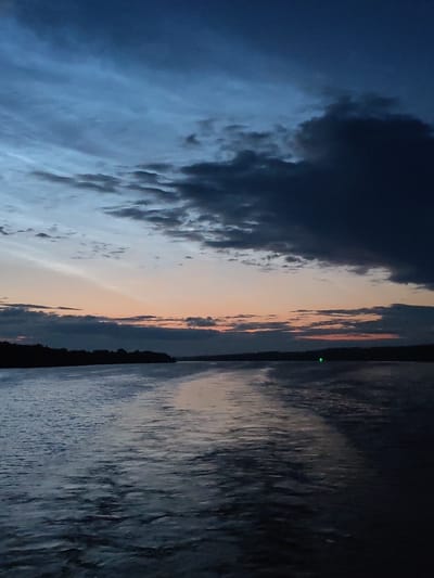 The image depicts a serene body of water, likely a river or lake, under a dramatic twilight sky. The sky transitions from a deep, dusky blue at the top to softer hues of orange and pink near the horizon, indicating either sunrise or sunset. Dark, heavy clouds dominate the upper portion of the sky, contrasting with lighter, streaky clouds lower down. A silhouetted tree line or landmass forms a dark band along the horizon. The water's surface is rippled, reflecting the muted colors of the sky and appearing dark and somewhat textured. A faint green light is visible on the horizon, possibly a buoy or distant navigational marker. The scene conveys a sense of calm and isolation.

While the image captures a natural landscape, it cannot be definitively placed in Ankimarovo, Russia. The provided image lacks any identifiable landmarks, signs, or features that would allow for such specific geographical localization.