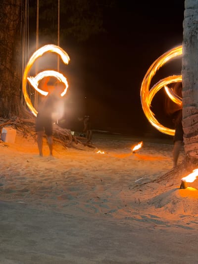 The image captures a nighttime fire show taking place on a sandy beach in Kamala, Thailand. Two individuals are actively engaged in fire spinning, creating bright orange and white light trails that form circular and arched patterns against the dark background. The performer on the left, wearing dark shorts and a top, stands barefoot on the light brown sand, illuminated by the fire. Behind this individual are exposed roots of a large tree and several vertical ropes or vines. A second performer is partially visible on the right, obscured by a thick tree trunk, also generating fire trails. The sand in the foreground and around the performers is brightly lit with an orange glow. Multiple small conical ground torches are positioned on the sand, emitting flames; one is visible in the lower right next to the tree trunk, and at least two more are further back. The dark expanse of the sea is visible in the background, and faint silhouettes of additional figures are present between the main performers. A white rectangular block lies on the sand to the left of the left performer.
