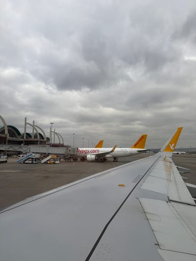This image depicts an airport apron scene under an overcast sky, viewed from an aircraft window overlooking its wing. The location is Sabiha Gökçen International Airport (SAW), situated near Tuzla, Istanbul, Turkey.

In the foreground, the light gray, metallic wing of a commercial airliner, from which the photograph is taken, extends across the lower right frame. The wing displays visible structural lines, panel seams, and numerous small, circular water droplets on its surface. A yellow rectangular safety marking is affixed to the wing.

In the midground, two white-bodied Pegasus Airlines aircraft are parked on the asphalt tarmac. The closer aircraft, with a yellow vertical stabilizer featuring a white Pegasus logo, displays "flypgs.com" in red and "PEGASUS" in yellow on its fuselage. This aircraft is connected to a passenger jet bridge that extends from the terminal building. Surrounding the aircraft are various ground support vehicles, including a blue and white mobile passenger stair unit, yellow baggage carts, and a small yellow tug vehicle. Several individuals, consistent with airport ground crew, are visible near the aircraft and jet bridge, engaged in operational activities. A second, identical Pegasus Airlines aircraft is parked further behind the first, partially obscured.

The background features the modern terminal building of Sabiha Gökçen International Airport, characterized by its distinctive arched support structures and extensive glass panels. Partially visible text on the terminal structure reads "INTERNATIONAL AIRPORT." Further in the distance, other aircraft tails and airport infrastructure are faintly visible. The sky above is entirely covered by dense, grey stratocumulus clouds. A tiny, distant aircraft is observable in the upper right quadrant of the sky.