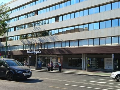 The image shows a street scene in front of El Corte Inglés department store in Sevilla, Spain. A dark colored car is driving on the road in the foreground. On the sidewalk, there is a bus stop with three people waiting. The El Corte Inglés building is visible behind the bus stop, with its distinctive signage. The building has a modern facade with rows of windows reflecting the blue sky. There are also advertisements visible on the side of the building. The scene appears to be a sunny day.