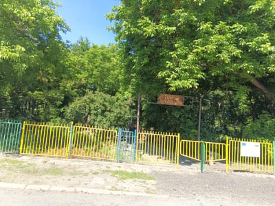 In the city of Tenevo, Bulgaria, a park entrance is depicted on a sunny day. A fence made of yellow and green vertical bars encloses the park. Behind the fence, lush green trees and foliage create a dense canopy. A brown wooden sign with Cyrillic text, likely indicating the park's name, is visible above the fence. A small blue gate is also present within the fence. A white sign with a European Union emblem and the Bulgarian flag is affixed to the right side of the yellow fence. The ground in front of the fence is a mixture of gravel and dirt.