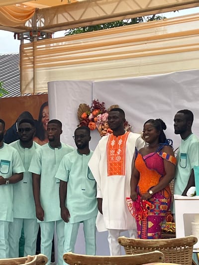 The image depicts a celebratory gathering in Kumasi, Ghana, featuring seven individuals positioned under a temporary canopy. At the center stands a man wearing a white tunic with orange embroidered patterns, next to a woman in a multi-colored off-shoulder dress predominantly featuring orange, pink, and blue patterns. Both individuals are smiling; the woman holds a red and orange fan-like object. Five other men surround the central pair; four are on the left and one on the right, all uniformly dressed in light green short-sleeved tunics with subtle darker green accents and matching trousers. Some of these men wear dark-rimmed glasses. Behind the central figures, a white backdrop is adorned with a large floral arrangement comprising maroon, peach, and gold-toned artificial flowers, interspersed with dried fan-shaped leaves. To the left, a large banner or poster partially displays the image of a man and a smiling woman. The scene is covered by an overhead structure consisting of metal trusses draped with light orange fabric. The foreground contains partially visible light-colored wooden furniture.