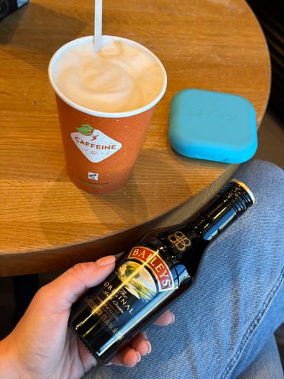An overhead, close-up shot depicts a casual indoor setting, likely a cafe, in Rīga, Latvia, during daylight hours. A person, visible by their legs clad in blue denim jeans and one hand, is seated at a light-colored wooden table.
The person's hand in the lower right holds a small, dark bottle of Baileys The Original Irish Cream liqueur. The label prominently displays "BAILEYS" and "The ORIGINAL Irish Cream," along with "PRODUCT OF IRELAND" and "50ml / 0.2L LIQUEUR 17% alc/vol: alc 17% vol."
On the wooden table, an orange-brown paper cup of coffee is present, featuring a frothy top and a white stir stick. The cup is branded with a lightning bolt graphic and the word "CAFFEINE," alongside "EST. 2007," "ROASTED COFFEE," and "ORGANIC." Next to the coffee cup, a light blue rectangular object with "SPARK" embossed on its surface is visible. The scene suggests a moment of leisure, possibly enjoying a coffee with an alcoholic addition.