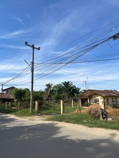 The image depicts a street scene in Kota Kuala Simpang, Indonesia, under a bright blue sky with wispy white clouds. The perspective is angled, with the majority of the frame dominated by a tangle of power lines crisscrossing against the sky. A wooden utility pole with multiple crossbars and insulators is visible, supporting these numerous wires.

To the right of the power lines, a dilapidated building with a weathered yellow facade and a rusty brown corrugated metal roof stands. Piles of wood and debris are gathered near the building, suggesting construction or disuse. Adjacent to the building is a dirt path and a paved road, with shadows from unseen objects falling across its surface, indicating it is daytime, likely morning or afternoon.

A grassy strip borders the road and the property, with a chain-link fence separating it from the more developed areas. Lush green trees, including palm trees, are scattered throughout the scene, adding to the tropical feel of the location. Further down the road, more residential structures with corrugated roofs are visible amidst the vegetation. No people are actively visible in the frame, and the overall impression is one of a quiet, somewhat rural or developing urban environment. The weather appears to be clear and sunny.