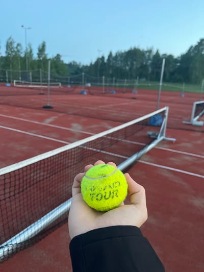 A first-person perspective shot shows a hand holding a neon yellow tennis ball on a red outdoor tennis court. The tennis ball has green lettering visible on its surface that reads "GRAND TOUR." The court has white lines marking boundaries, and a tennis net stretches across the foreground. In the background, more tennis courts are visible, along with trees and lampposts, suggesting a sports complex. The sky is a clear, pale blue, indicating it is likely early morning or late evening. The scene appears to be outdoors, possibly at a tennis club or park, and the setting suggests a location in Vantaa, Finland, given the commonality of outdoor sports facilities in the region.