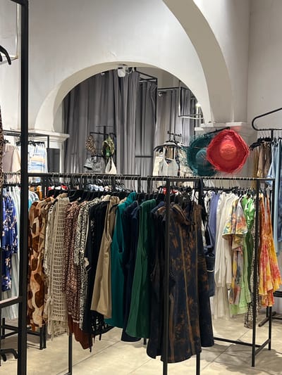 This is a vertical shot of a clothing store display. The store is brightly lit, suggesting it's daytime. The display features racks of neatly folded clothes, predominantly in earthy tones and vibrant patterns. Among the folded garments, there are also several hats visible, one a bright coral color with feathers, and another a deep teal. The store's interior includes a distinctive white arched wall and grey curtains in the background, hinting at a modern yet possibly slightly dated design. The overall impression is that of a retail environment showcasing a variety of apparel, possibly a boutique given the unique hat selection and patterned fabrics. There is no visible text in the image.