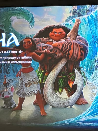 This media file features a promotional image, likely a movie poster or cover art, for the animated film "Moana." The central subjects are the characters Moana, a young Polynesian woman, and Maui, a demigod. Moana is depicted with dark hair and a warm expression, wearing a traditional island skirt and necklace. Maui is larger and more imposing, adorned with intricate tribal tattoos and holding a magical fishhook.

The scene is set against a backdrop of a dramatic, swirling blue wave, characteristic of the film's oceanic theme, and a sandy beach. Lush green foliage is visible, intermingled with Maui's hook, suggesting a tropical island environment. A small, pink pig, likely the character Pua, is also visible at the bottom right of the frame, adding a touch of whimsy.

The visible text on the poster is in Russian. It indicates "1 ч 43 мин." (1 hour 43 minutes) which is the movie's runtime, and "6+" which signifies a general audience rating suitable for viewers aged 6 and above. The other text, "от природу от гибели. НЯМИ И ИСПытаниями", translates to something like "from nature from perishing. WITH trials and tribulations," hinting at the film's plot involving a quest to save their homeland. The overall mood is adventurous and heroic, with the characters poised for action against the powerful forces of nature. The image is likely a still captured on a screen, given its slightly blurry quality and the visible screen texture.