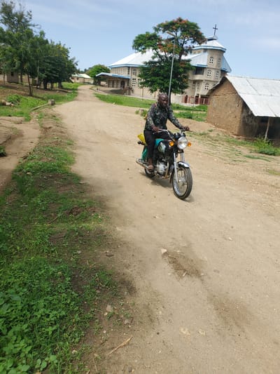 A man is pictured riding a motorcycle on a dirt road in a village setting. He is wearing a camouflage shirt and dark pants, with a yellow bag slung over his shoulder. The motorcycle is a standard street bike, with a chrome headlight and orange turn signals. The road is unpaved and dusty, with patches of green grass growing on the sides. In the background, there is a large building that appears to be a church, with a white roof and several windows. The building has a cross on top, indicating its religious affiliation. There are also trees and other vegetation visible in the background, suggesting a rural or semi-rural location. The sky is blue and clear, indicating a daytime scene with fair weather. The man appears to be looking at the camera, with a neutral expression on his face. The overall scene is one of everyday life in a Nigerian village, with a focus on the man and his motorcycle. The image captures a moment of transit, as the man makes his way along the dirt road. The presence of the church suggests a community setting, where religious institutions play a prominent role.