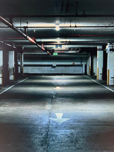 The image depicts an empty, dimly lit parking garage. The perspective is from ground level, looking down a long, straight aisle. The ceiling is lined with pipes and fluorescent lights, casting shadows on the concrete floor. White lines demarcate parking spaces, and a large white arrow painted on the ground indicates the direction of traffic. The walls are a light, textured material, with vertical yellow protective poles at intervals. There are no people or vehicles visible in the scene. The overall atmosphere is industrial and stark. The image does not contain any information or visual cues that would allow for its location to be identified as Kursk, Russia.